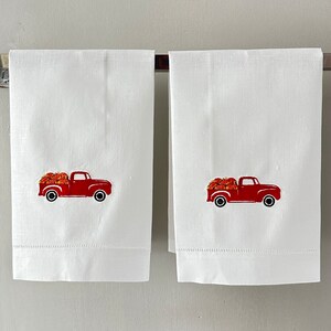 Op de afbeelding: Twee witte handdoeken met rode geborduurde afbeeldingen van een pick-up truck met pompoenen.