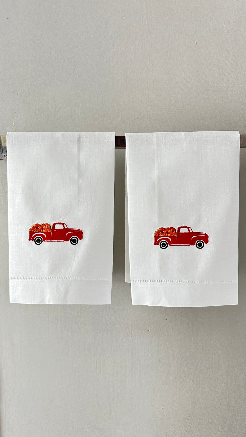 Op de afbeelding: Twee witte handdoeken met rode geborduurde afbeeldingen van een pick-up truck met pompoenen.