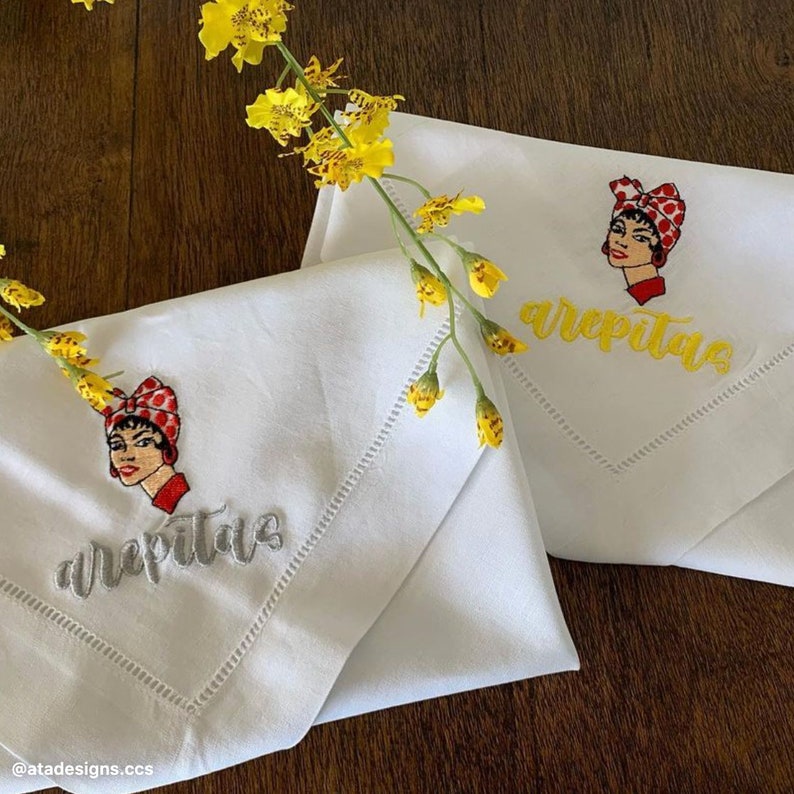 May include: Two white linen napkins with embroidered images of a woman with a red and white polka dot headscarf. The napkins are folded in half and the embroidery says "arepitas". One napkin has yellow embroidery and the other has silver embroidery.