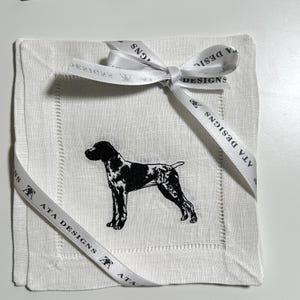 May include: White linen cocktail napkin with a black and white dog illustration. The napkin is tied with a white ribbon that has "ATA DESIGNS" printed on it. The napkin has a stitched border.