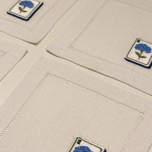 May include: A set of square off-white linen napkins featuring a hemstitched border and an embroidered patch in one corner. The patch displays a blue hydrangea flower with green leaves and the number 2 in dark blue thread on a white background.