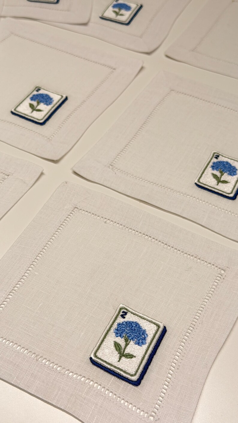 May include: A set of square off-white linen napkins featuring a hemstitched border and an embroidered patch in one corner. The patch displays a blue hydrangea flower with green leaves and the number 2 in dark blue thread on a white background.