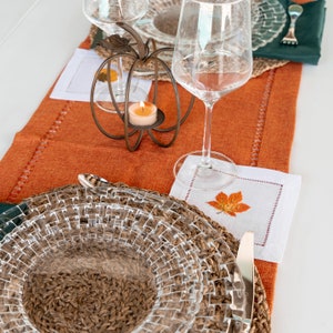 May include: A close-up of a table setting with a woven placemat, a clear glass plate, a clear glass wine glass, a candle in a glass holder, and a white napkin with an embroidered leaf design.