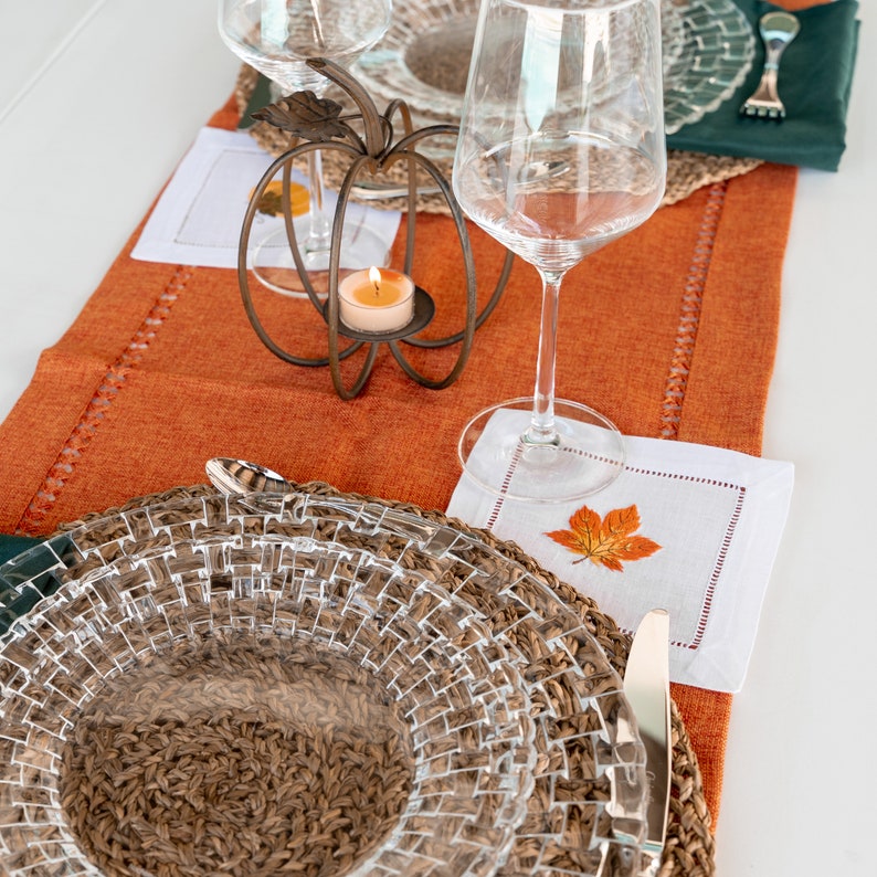 May include: A close-up of a table setting with a woven placemat, a clear glass plate, a clear glass wine glass, a candle in a glass holder, and a white napkin with an embroidered leaf design.