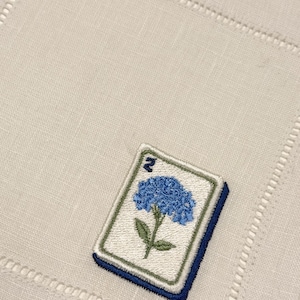 May include: A square off-white linen napkin featuring a hemstitched border and an embroidered mahjong tile motif. The tile displays a blue hydrangea flower with green leaves and the number two in the upper corner, rendered in blue thread.