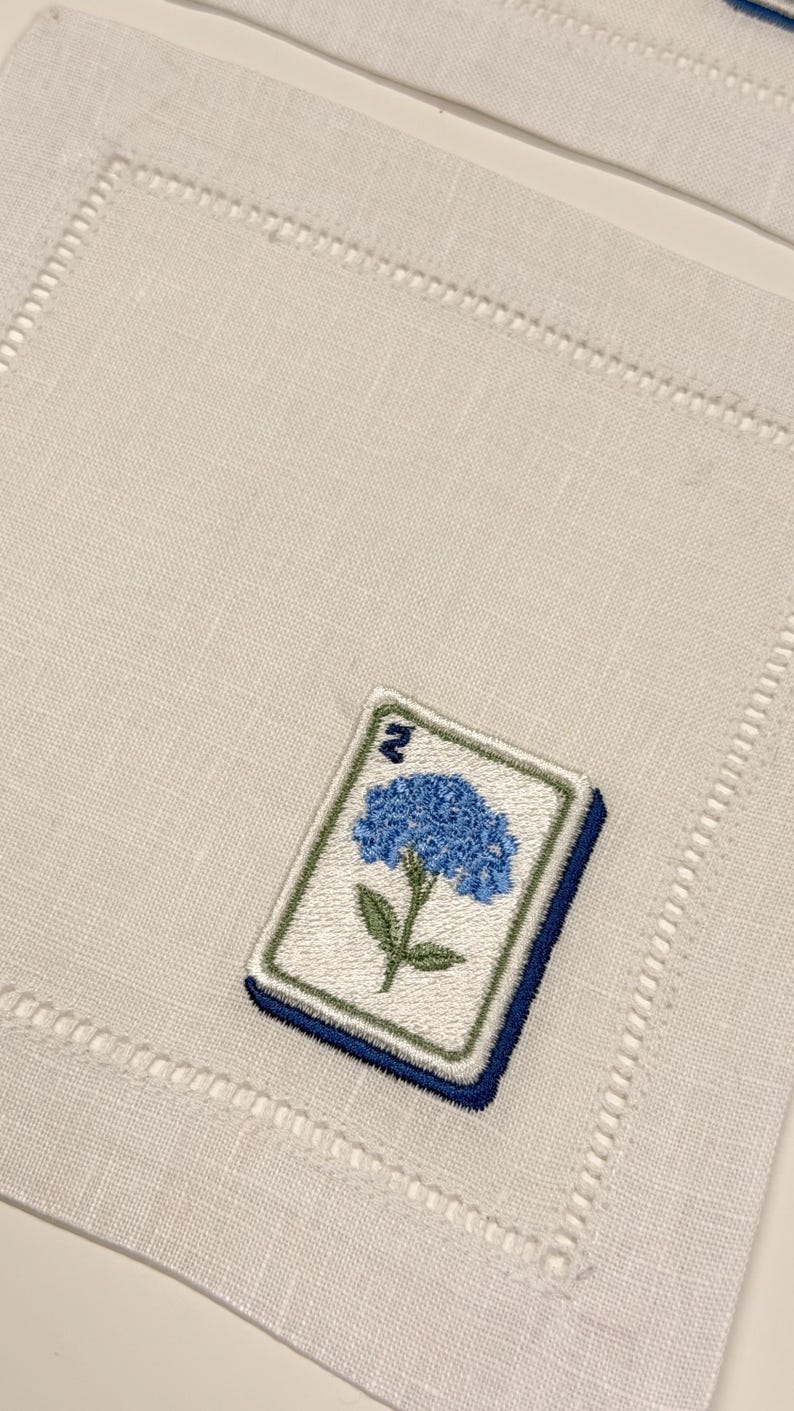 May include: A square off-white linen napkin featuring a hemstitched border and an embroidered mahjong tile motif. The tile displays a blue hydrangea flower with green leaves and the number two in the upper corner, rendered in blue thread.