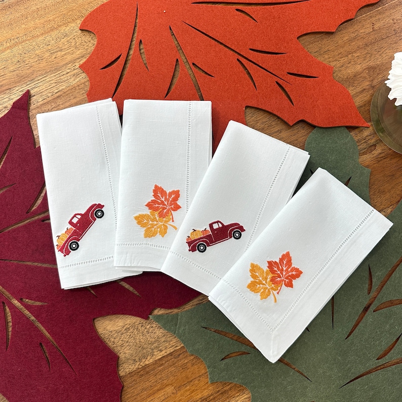 Op de afbeelding: Vier witte linnen servetten met geborduurde herfstthema's. Elk servet heeft een rode vrachtwagen met pompoenen en een groep oranje en gele bladeren.