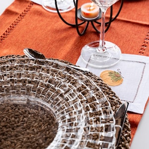 May include: A clear glass plate with a woven brown placemat underneath. A silver fork and knife are on the placemat. A white napkin with an embroidered pumpkin design is on the table.