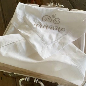 May include: White linen napkin with a brown embroidered design of three pancakes and the word "crepes" written in cursive.