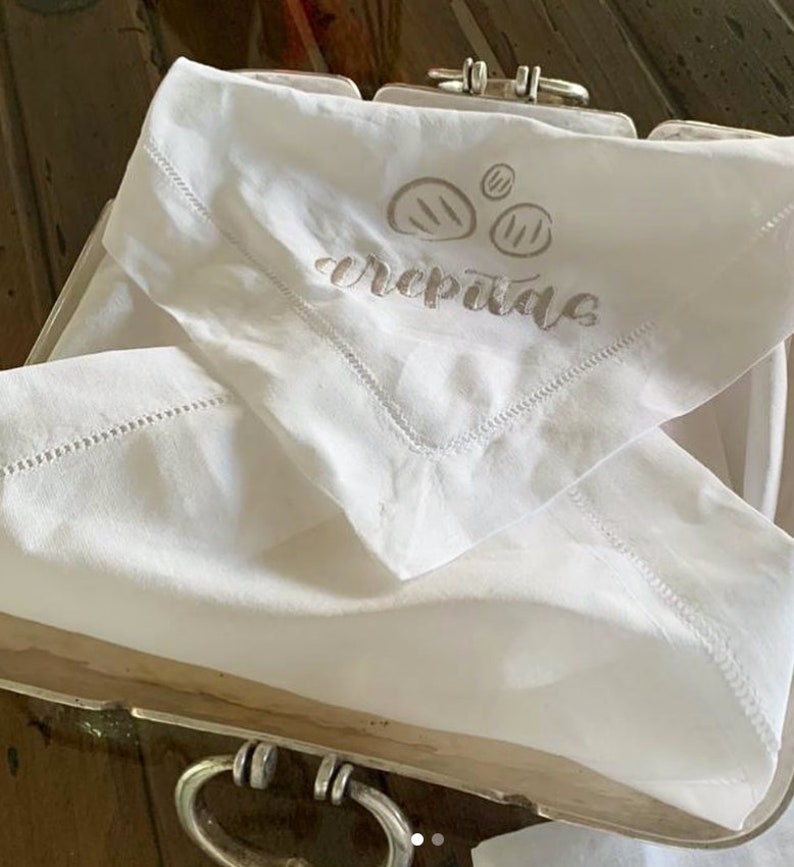 May include: White linen napkin with a brown embroidered design of three pancakes and the word "crepes" written in cursive.