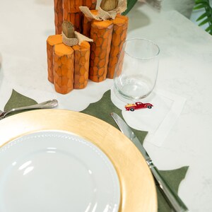 May include: A gold charger plate with a white dinner plate on top. A green and brown placemat with a white napkin with a red truck design is on the table. There are orange and brown wooden pumpkins on the table.