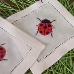 May include: White linen napkins with a red and black ladybug design. The napkins have a stitched border and are placed on a green surface. The ladybugs have black spots and are embroidered.