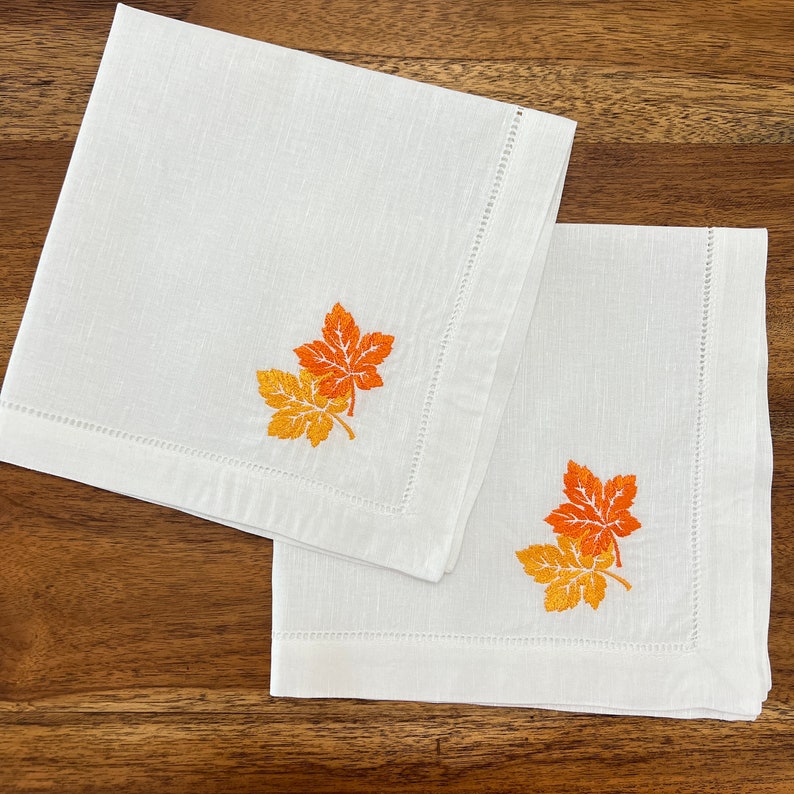 Op de afbeelding: Twee witte linnen servetten met geborduurde oranje en gele herfstbladeren.