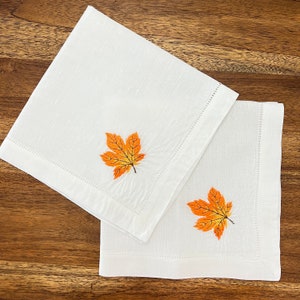 Op de afbeelding: Twee witte linnen servetten met oranje en gele geborduurde herfstbladeren.
