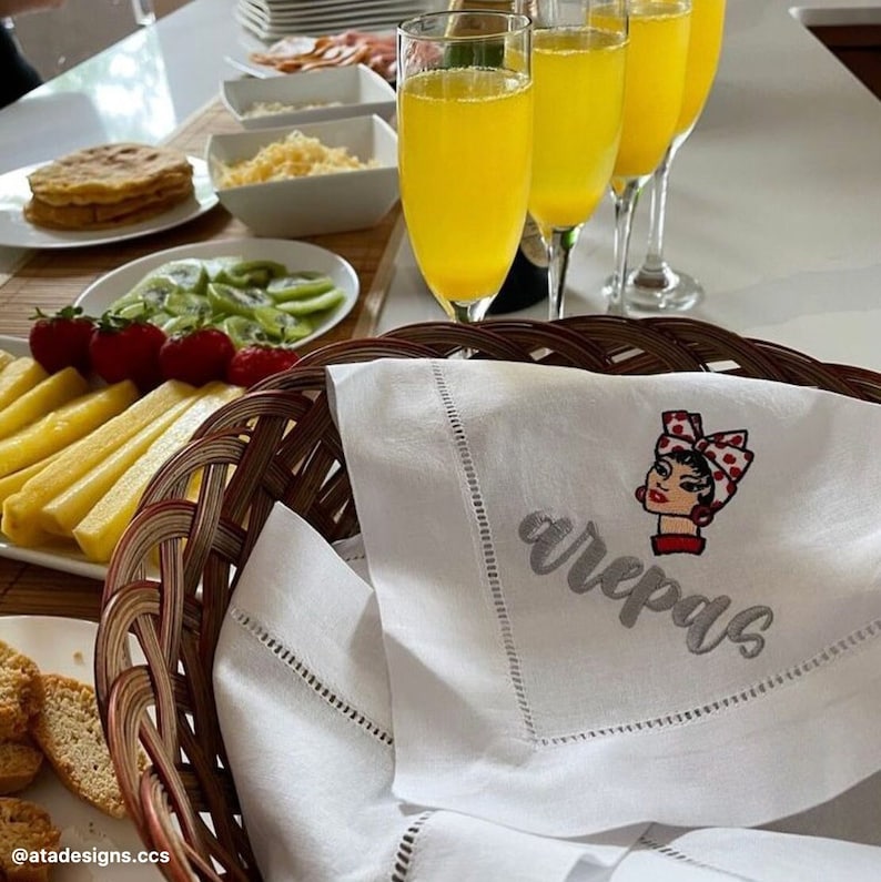 May include: White linen napkin with embroidered text "arepas" and a cartoon image of a woman with a red and white polka dot headband. The napkin is folded and placed in a wicker basket.