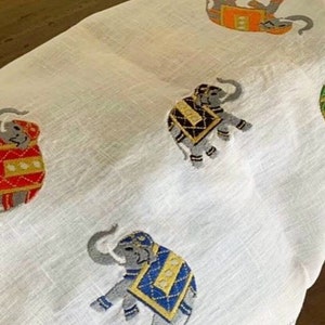 Puede incluir: Tela blanca con elefantes bordados en varios colores, incluyendo rojo, azul, verde y negro. Los elefantes llevan sillas de montar decorativas y tienen detalles intrincados.