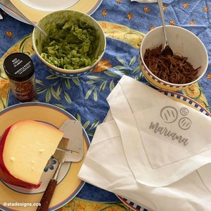 May include: A white napkin with the word "mariana" embroidered on it, a bowl of shredded meat, a bowl of guacamole, a wedge of red cheese on a yellow plate, and a small jar of red chili flakes.