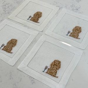 May include: Set of four white linen cocktail napkins with embroidered brown dogs holding a champagne glass.