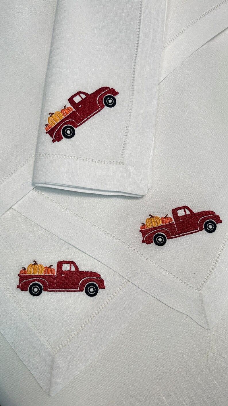 Op de afbeelding: Drie witte linnen servetten met rode geborduurde vrachtwagens die pompoenen vervoeren. De vrachtwagens zijn naar rechts gericht en hebben een rode cabine en een witte laadbak. De pompoenen zijn oranje en geel.