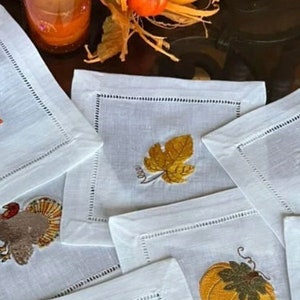 May include: Set of six white linen napkins with embroidered fall-themed designs. The designs include a pumpkin, a turkey, a leaf, and a cornucopia.