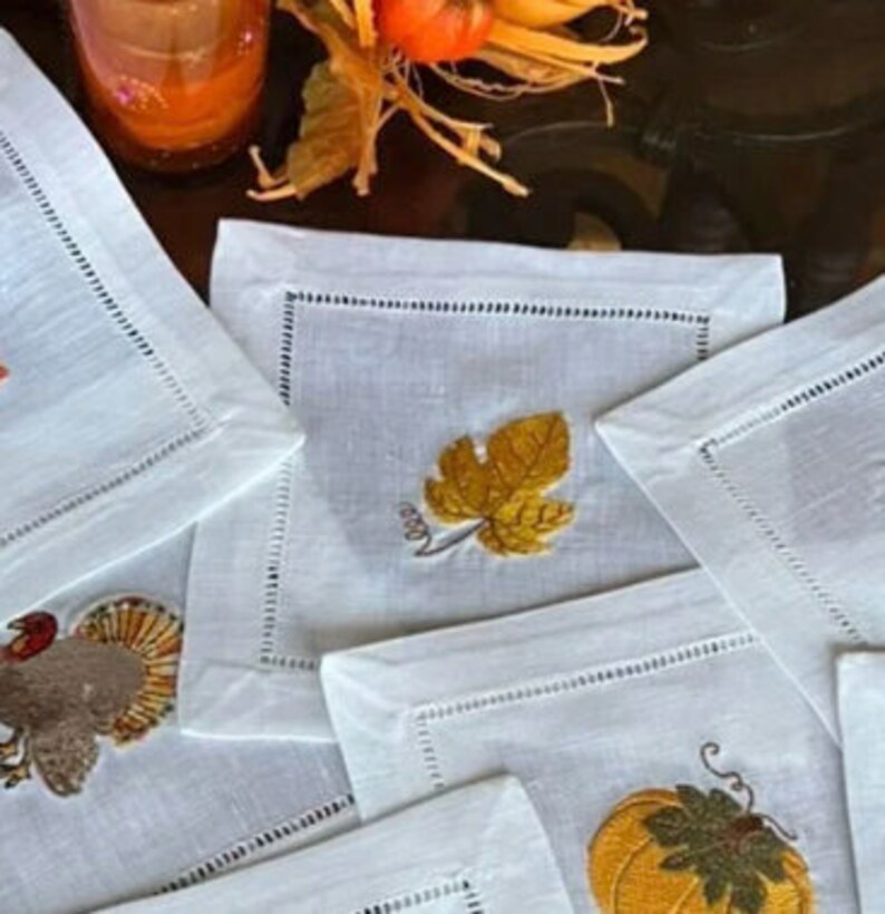 May include: Set of six white linen napkins with embroidered fall-themed designs. The designs include a pumpkin, a turkey, a leaf, and a cornucopia.