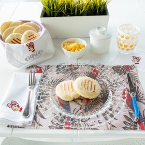 May include: A white table setting with a clear glass plate, two arepas, a white napkin with a red and white illustration, a white napkin with the word "arepas" embroidered on it, a white ceramic sugar bowl, a glass of orange juice, and a white ceramic bowl with shredded cheese.