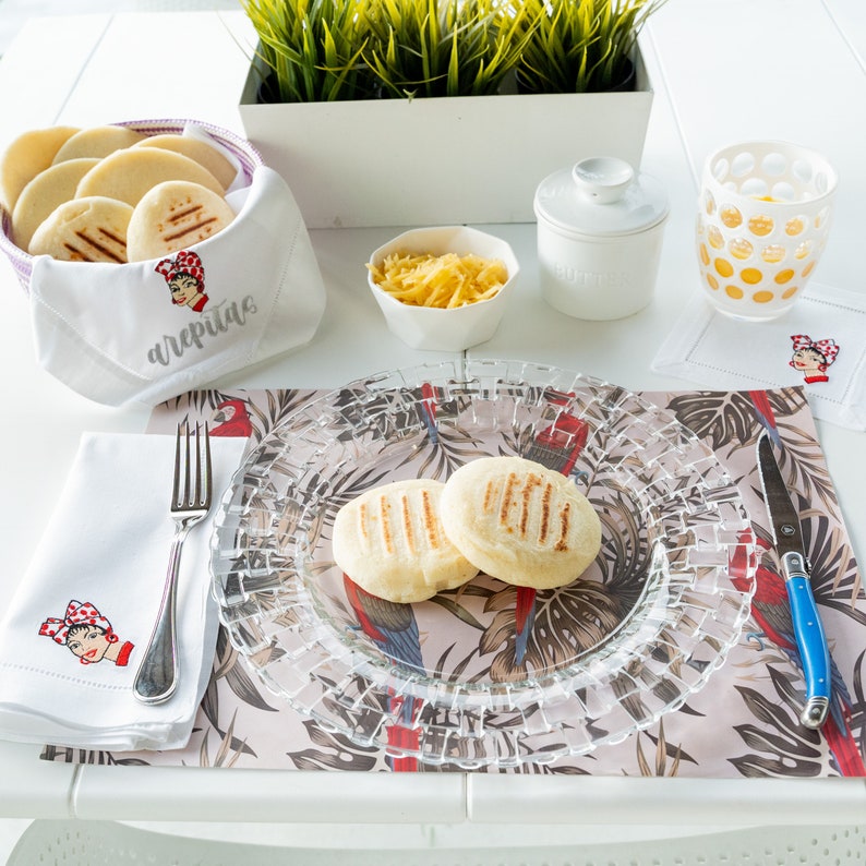 May include: A white table setting with a clear glass plate, two arepas, a white napkin with a red and white illustration, a white napkin with the word "arepas" embroidered on it, a white ceramic sugar bowl, a glass of orange juice, and a white ceramic bowl with shredded cheese.