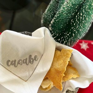 May include: A white linen napkin with a gray embroidered word "casabe" on it. The napkin is folded and placed on a table with a pile of crispy, golden brown flatbread on top.