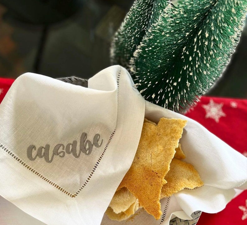 May include: A white linen napkin with a gray embroidered word "casabe" on it. The napkin is folded and placed on a table with a pile of crispy, golden brown flatbread on top.