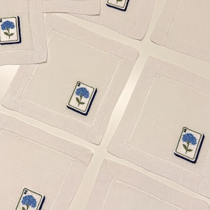 May include: A set of square white linen napkins featuring a hemstitched border and an embroidered patch of a blue hydrangea flower above the number 2, designed to resemble a mahjong tile with a dark blue border on each individual napkin.