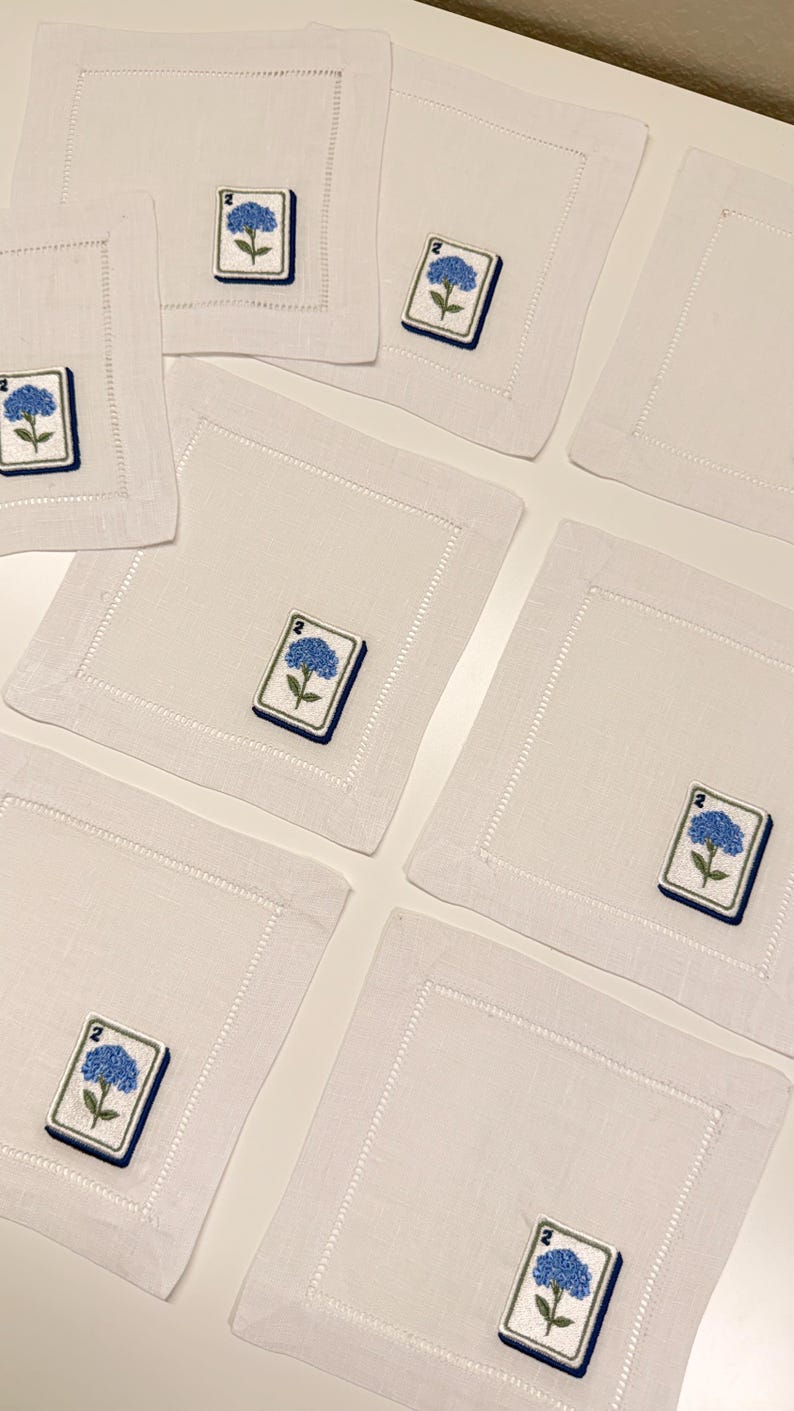 May include: A set of square white linen napkins featuring a hemstitched border and an embroidered patch of a blue hydrangea flower above the number 2, designed to resemble a mahjong tile with a dark blue border on each individual napkin.