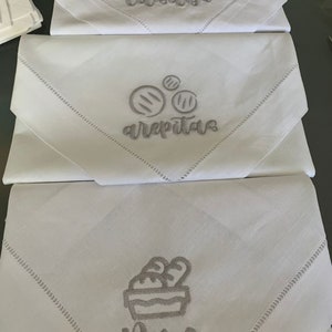 May include: Three white linen napkins with embroidered designs. The top napkin says "casaba", the middle napkin says "arepitas" with a drawing of three arepitas, and the bottom napkin says "pan" with a drawing of a basket of bread.