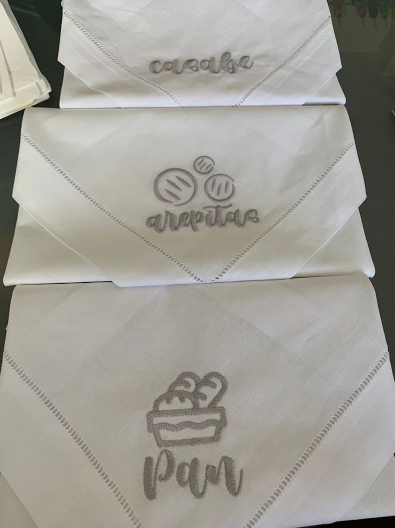 May include: Three white linen napkins with embroidered designs. The top napkin says "casaba", the middle napkin says "arepitas" with a drawing of three arepitas, and the bottom napkin says "pan" with a drawing of a basket of bread.