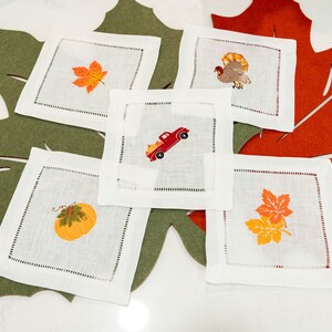 Op de afbeelding: Set van zes witte linnen servetten met geborduurde herfstthema-ontwerpen. De ontwerpen omvatten een pompoen, een vrachtwagen met pompoenen, een kalkoen en twee verschillende bladerenontwerpen. De servetten zijn gerangschikt op een groene en oranje viltbladerenachtergrond.