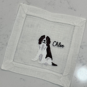 Könnte beinhalten: Weiße Leinen-Serviette mit gesticktem braunen und weißen Hund und dem Namen Chloe.
