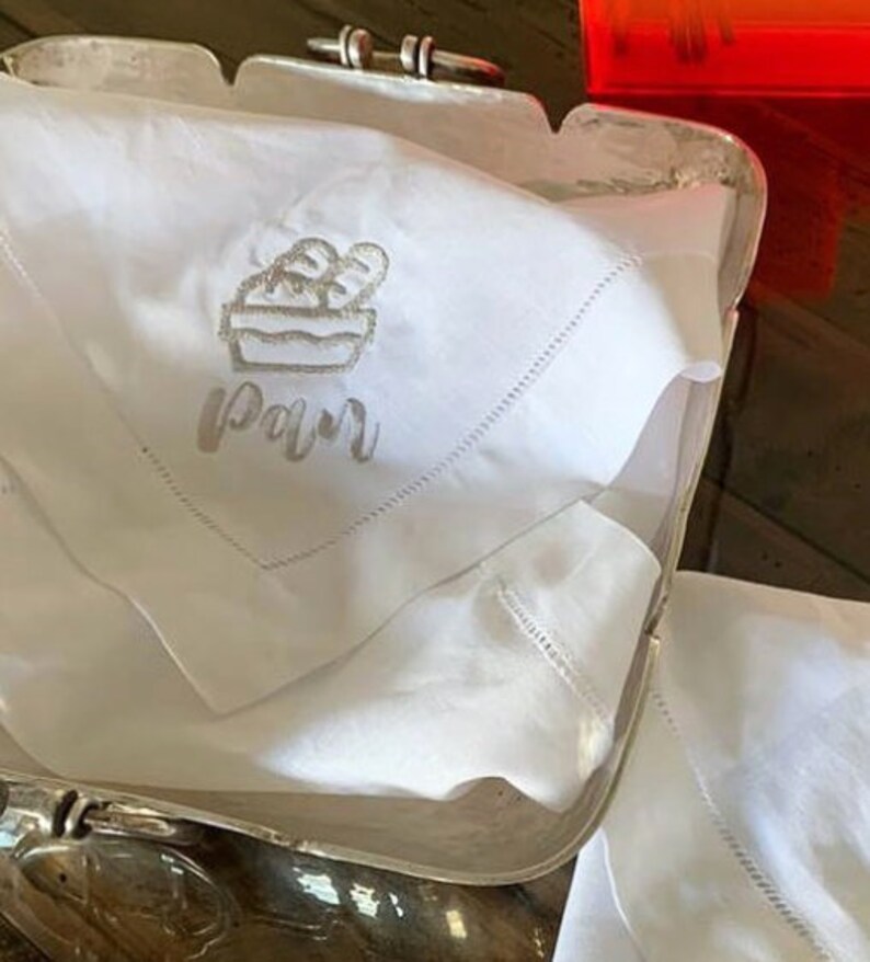 May include: White linen napkin with embroidered bread basket and the word "Pain" in cursive.