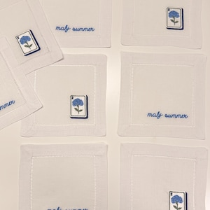 May include: A set of six square white linen napkins featuring embroidered details. Some napkins display the blue text "maly summer" in cursive, while others feature an embroidered blue hydrangea flower on a white rectangular tile with the number 2.