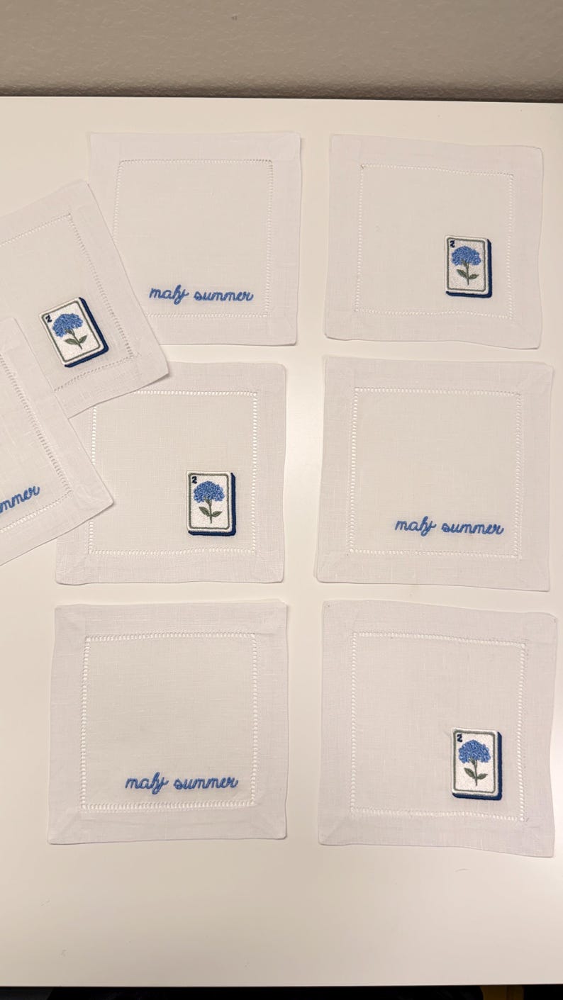 May include: A set of six square white linen napkins featuring embroidered details. Some napkins display the blue text "maly summer" in cursive, while others feature an embroidered blue hydrangea flower on a white rectangular tile with the number 2.