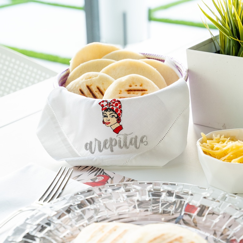 May include: A white napkin with embroidered text "arepitas" and a cartoon image of a woman wearing a red and white polka dot headscarf. A basket filled with grilled arepas is sitting on the napkin.