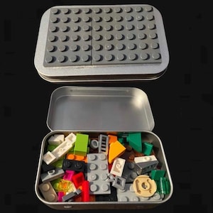 May include: A silver metal tin with a lid that has a gray Lego baseplate on the inside. The tin is open and filled with a variety of colorful Lego bricks.