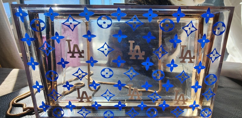 May include: A clear plastic box purse with a gold chain strap. The purse is decorated with a repeating pattern of blue four-leaf clover and brown diamond shapes. The letters "LA" are repeated in gold on the purse.