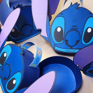 May include: Four blue and purple papercraft boxes shaped like Stitch from the Disney movie Lilo & Stitch. Each box has a handle and a pair of ears.