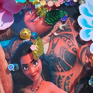 May include: A close-up of a handmade craft featuring a photo of Moana and Maui from the Disney movie Moana. The photo is surrounded by colorful embellishments, including flowers, leaves, and glitter. The craft is set against a green background.