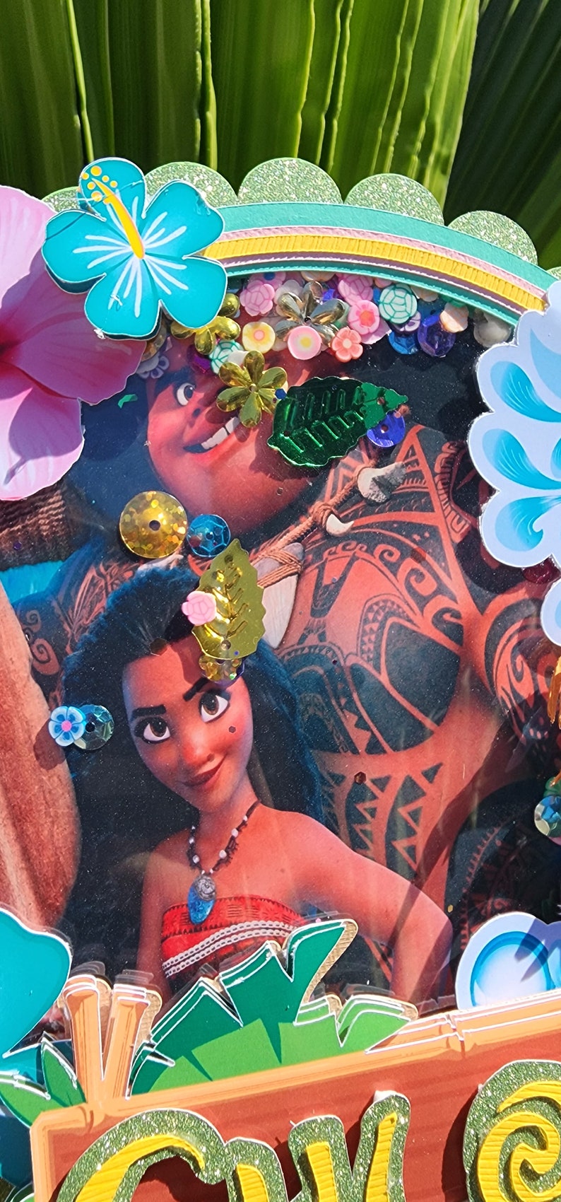 May include: A close-up of a handmade craft featuring a photo of Moana and Maui from the Disney movie Moana. The photo is surrounded by colorful embellishments, including flowers, leaves, and glitter. The craft is set against a green background.