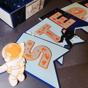May include: An astronaut-shaped light with an orange illuminated helmet and a banner spelling out "STEP" with space-themed illustrations. The letters are gray with orange accents, and the banner is light blue with a dark blue border.