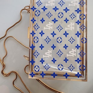 May include: A clear acrylic box clutch with a gold chain strap. The box has a white and blue pattern with the letters "LA" repeated throughout.