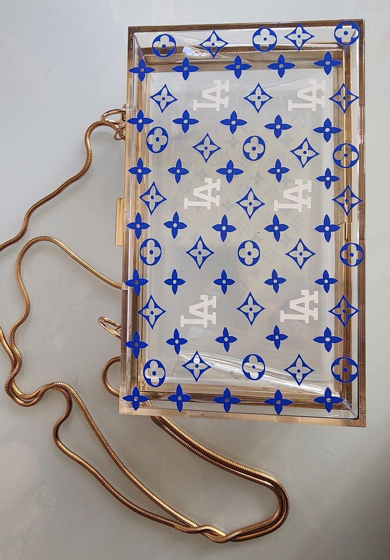 May include: A clear acrylic box clutch with a gold chain strap. The box has a white and blue pattern with the letters "LA" repeated throughout.