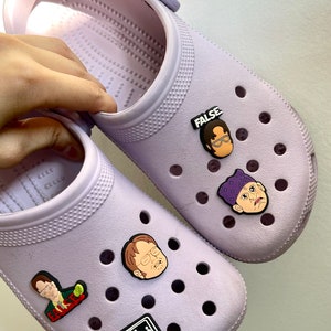 May include: A pair of lavender clogs with various "The Office" themed Jibbitz charms. The charms include characters like Dwight Schrute, Michael Scott, and Jim Halpert, as well as the text "The Office" and "False".