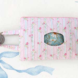 Peut inclure: Une pochette en tissu rectangulaire avec un motif à rayures roses et blanches et de petites fleurs. La pochette présente une ouverture ovale centrale révélant un intérieur à fleurs bleues, une fermeture éclair rose et une poignée à deux sphères.