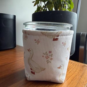 May include: A glass jar encased in a fabric sleeve featuring a pattern of white geese with orange beaks and feet, interspersed with small pink floral clusters on a background of thin, vertical beige and white stripes.